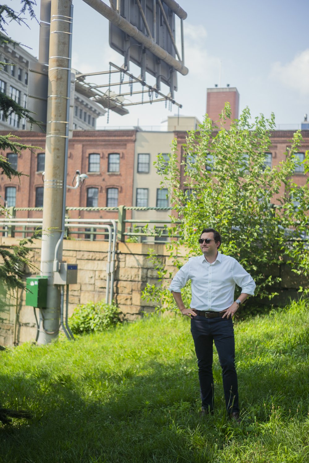 A man wearing a plain white button down shirt and dark pants stands in a grassy area with an elevated road sign a few feet behind him
