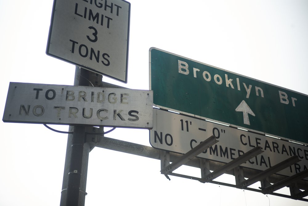 Four road signs, three are white with black lettering and one is green with white lettering. They read "Brooklyn Br" "To Bridge No Trucks" "Weight Limit 3 Tons" and "11 feet clearance No Commercial Traffic"