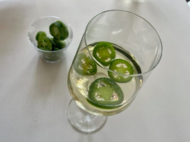 A wine glass with a plastic cup of frozen jalapenos beside it, and sliced jalapenos in the glass.