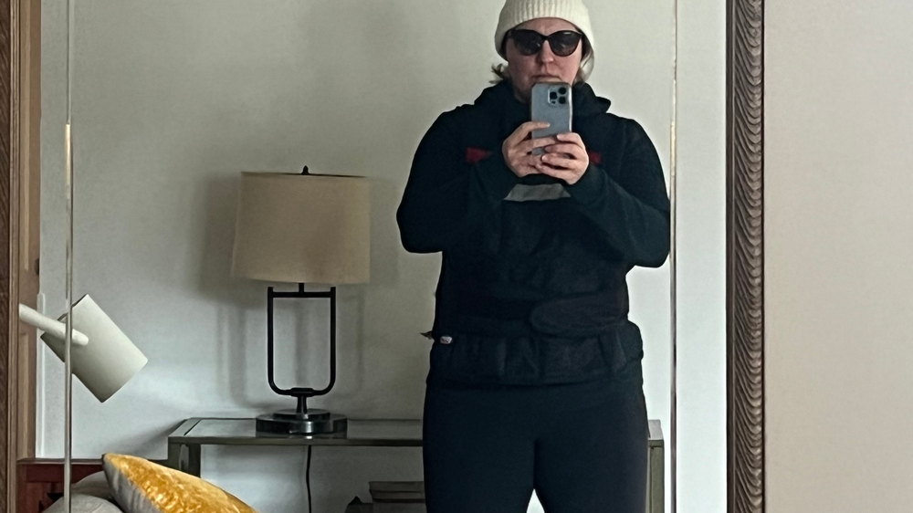 A woman poses in the mirror wearing all black and a white beanie.