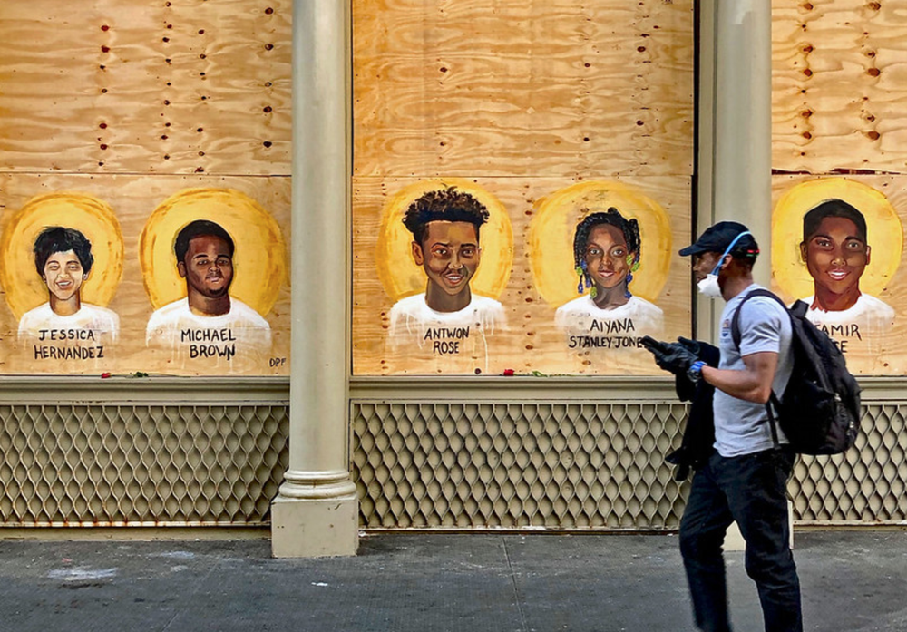 A photo of a person walking by a boarded up store in SoHo featuring the faces of people killed by police