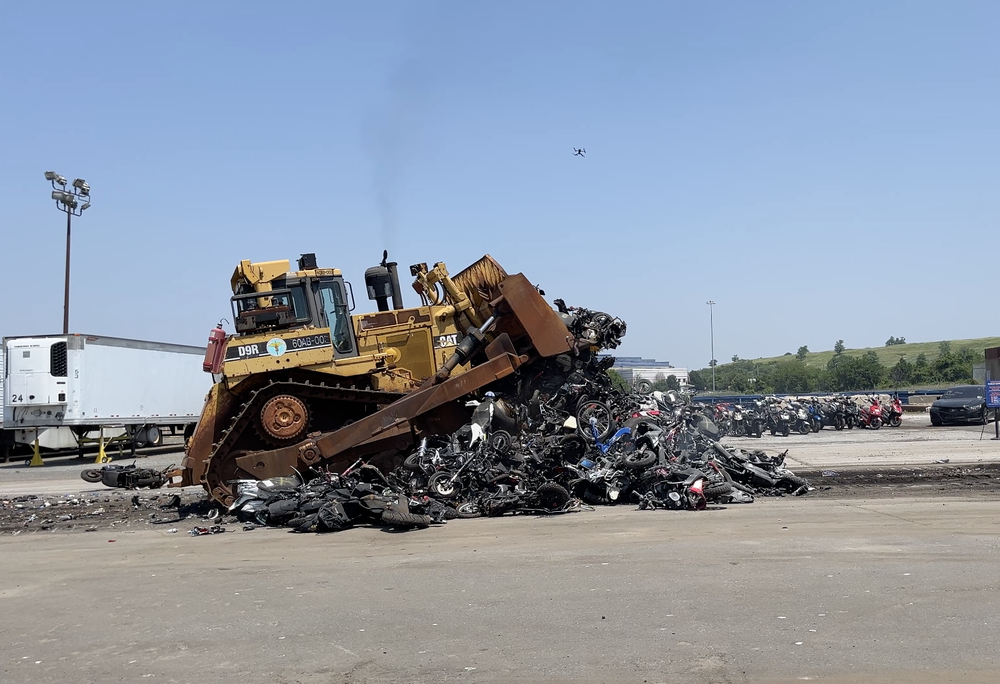A bulldozer runs over a pile of e-bikes and mopeds while a little drone flied overhead.