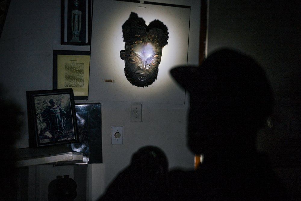 In a dark room, a flashlight illuminates an African mask that hangs on a wall.