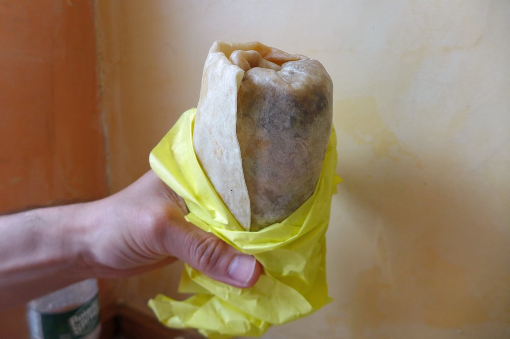 A person holds a burrito in a yellow tissue paper.