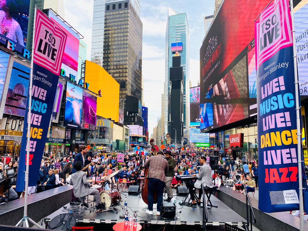 Musicians playing on an outdoor stage, viewed from behind with the audience visible beyond them.