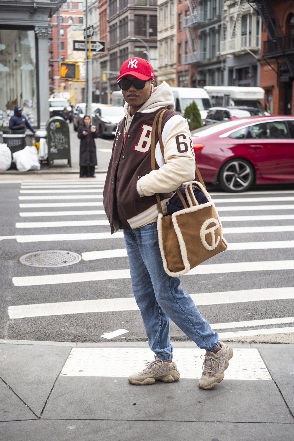 A man poses on a New York City sidewalk wearing a brown bomber jacket.