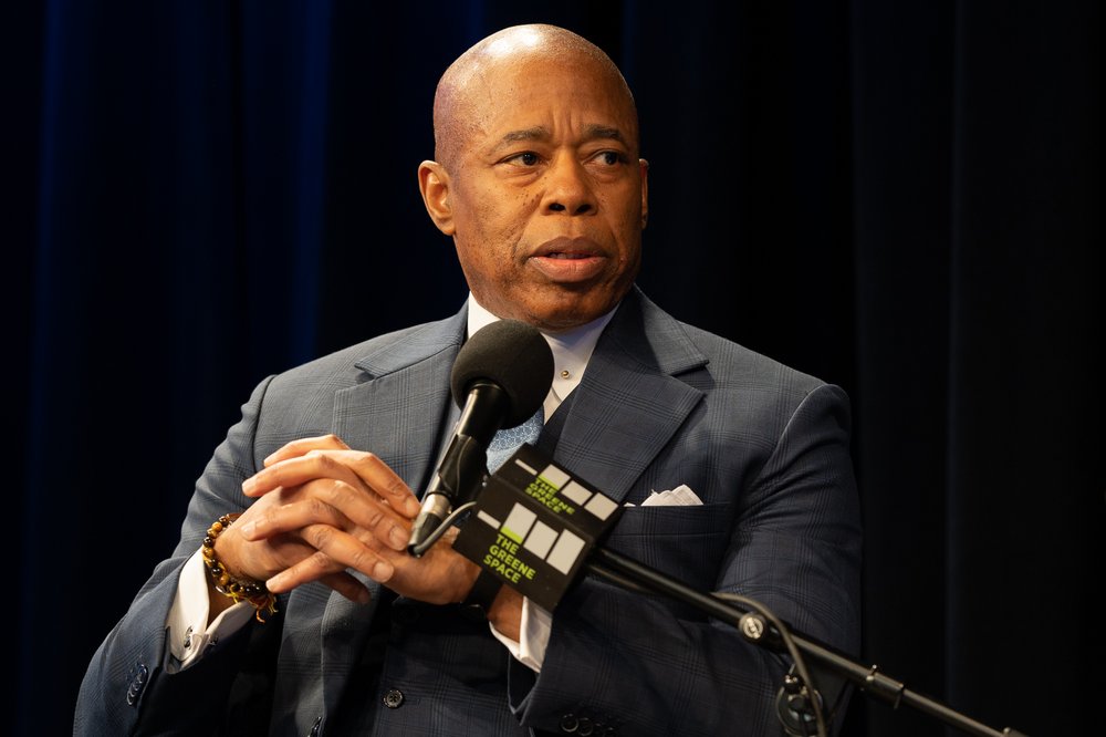 Mayor Eric Adams in a blue plaid suit, clasps his hands while talking into a microphone.