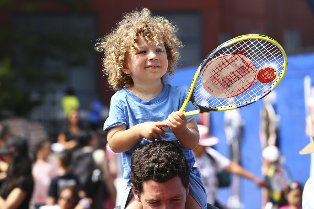 A young child holding a tennis racket sits on a grownup's shoulders.