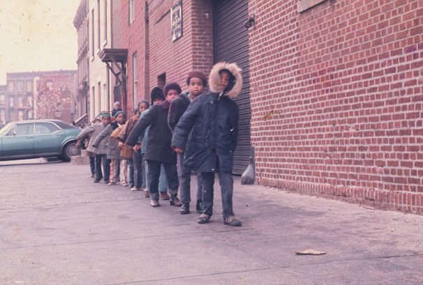 A vintage photograph of  a large group of children milling about on the sidewalk in front of a community center.