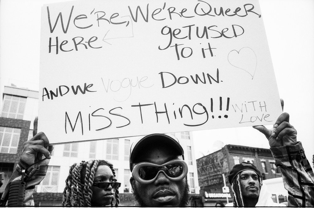 A person holds a sign that reads: "We're here, we're Queer, get used to it. And we vogue down Ms. Thing!!! With love."