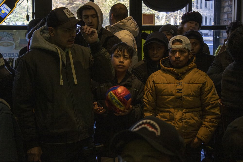 A crowd of soccer fans stand shoulder to shoulder in a bar, watching a match. A child in the center holds a multi-colored soccer ball.