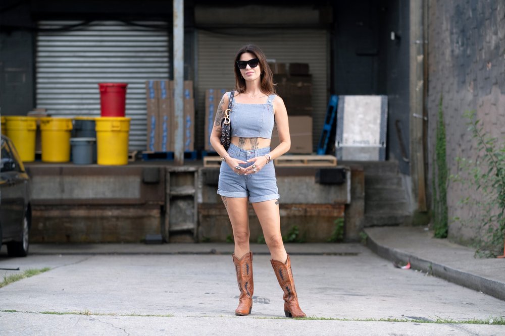 A woman wears a blue tank top, shorts, tan leather boots and sunglasses.