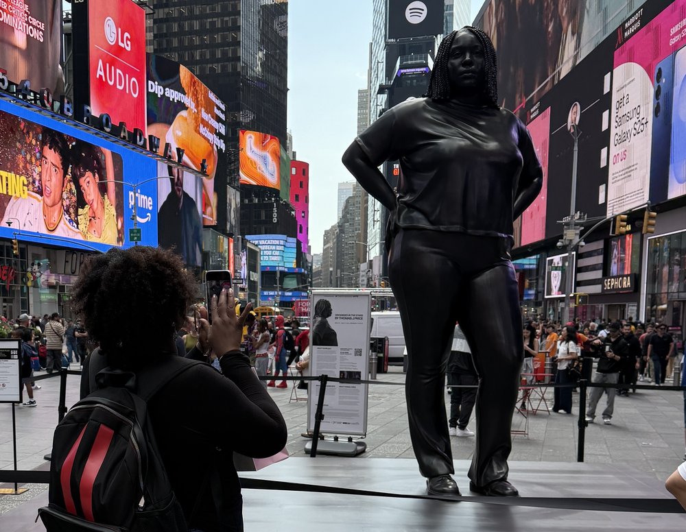 A woman with her back to the camera takes a picture of a statue in Times Square.