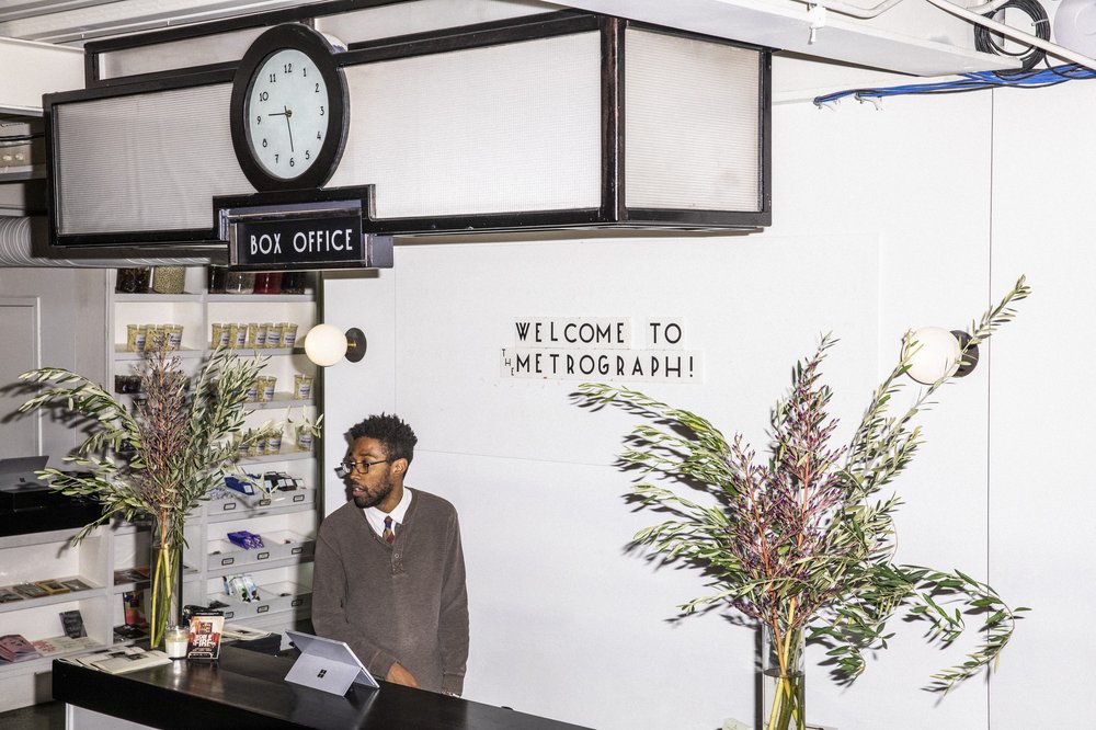 A man stands behind the counter of the Metrograph movie theater. A sign behind him reads "Welcome to Metrograph."