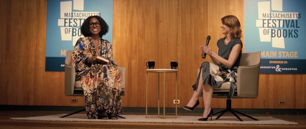 Two women sit in chairs on a stage in front of an audience.
