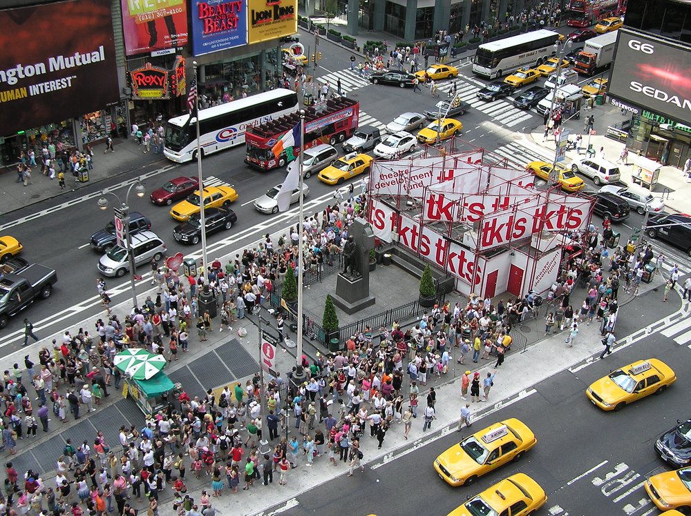 An overhead view of a crowded Times Square, with a booth topped by white signs at one end.