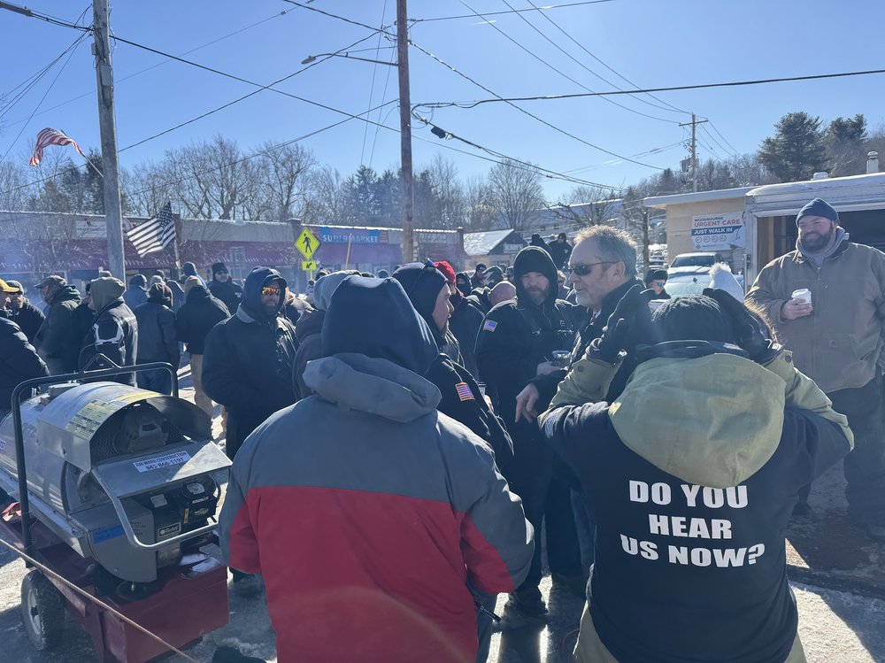 A group of people stand outside wearing winter gear. A person with their back to the camera wears a shirt reading DO YOU HEAR US NOW?