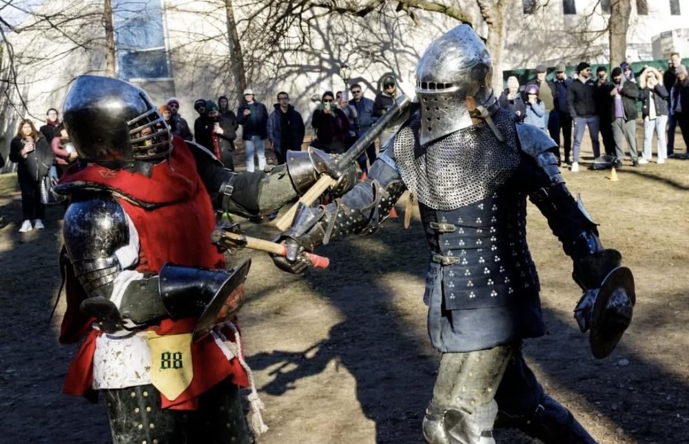 Two people fight outdoors in medieval armor as a crowd in modern-day clothing looks on.