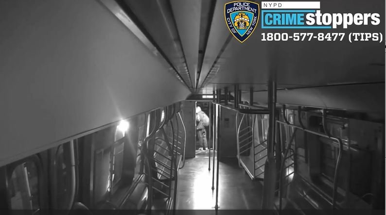 A still from a surveillance video showing a person wearing a hood on an empty, unlit subway car.