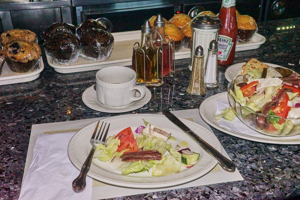 A plate of Greek salad is seen next to a serving bowl of Greek salad. The dishes are on top of a diner counter with oil, vinegar, salt, pepper, sugar, a bottle of ketchup, and wrapped muffins in the background.