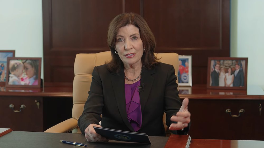 Governor Kathy Hochul sits at a table with a hand up answering a question