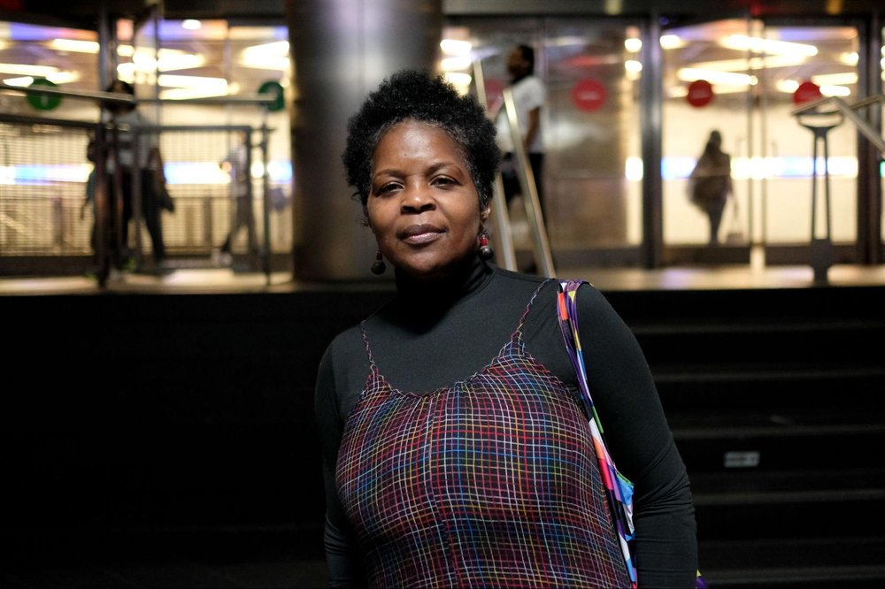 A woman in a red plaid dress and black turtleneck stands on a subway platform