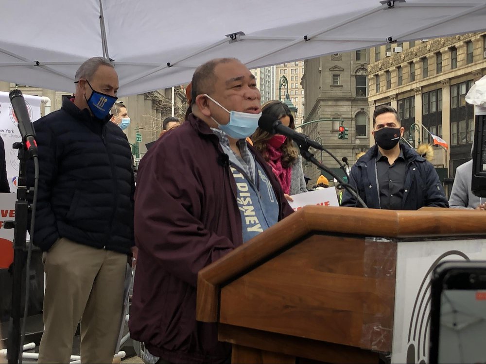 Noel Quintana speaks at the lectern, with his mask lowered to show the scar across his face