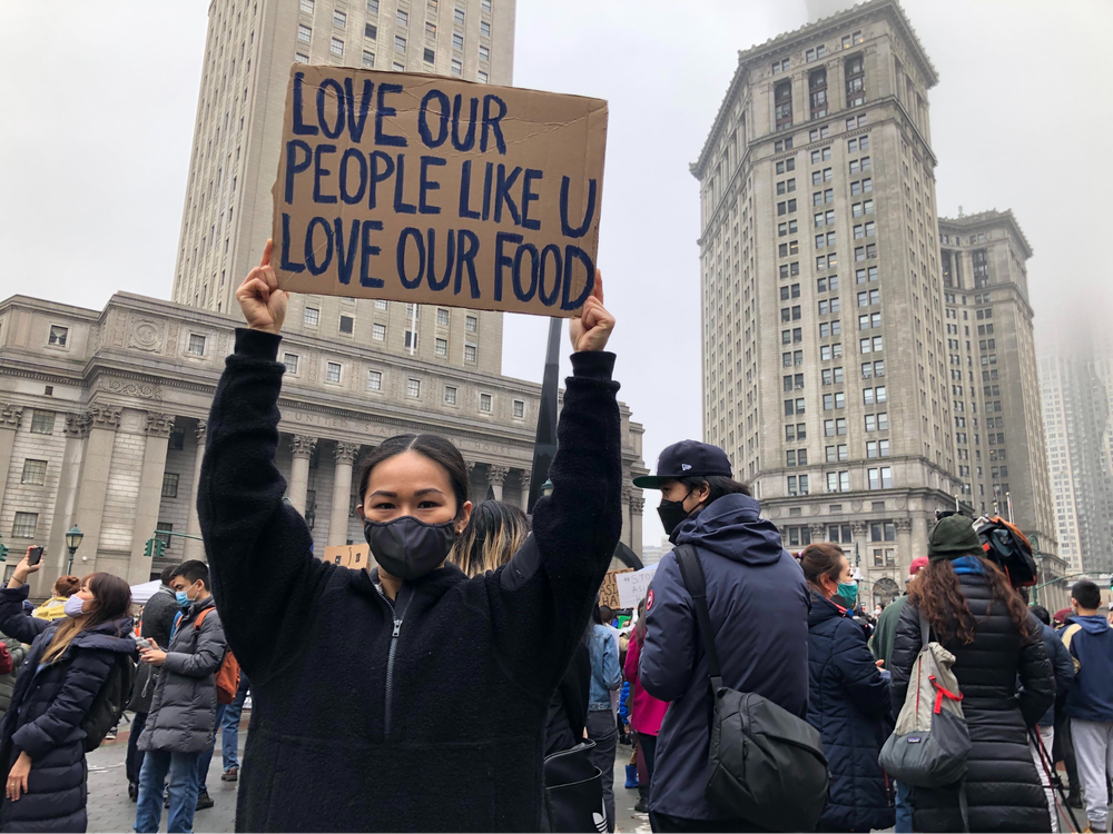 A woman holds a sign that says "Love Our People Like U Love Our Food"