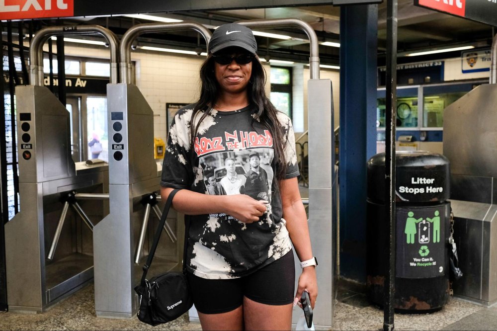 A woman wearing a baseball cap and sunglasses stands in front of subway turnstiles