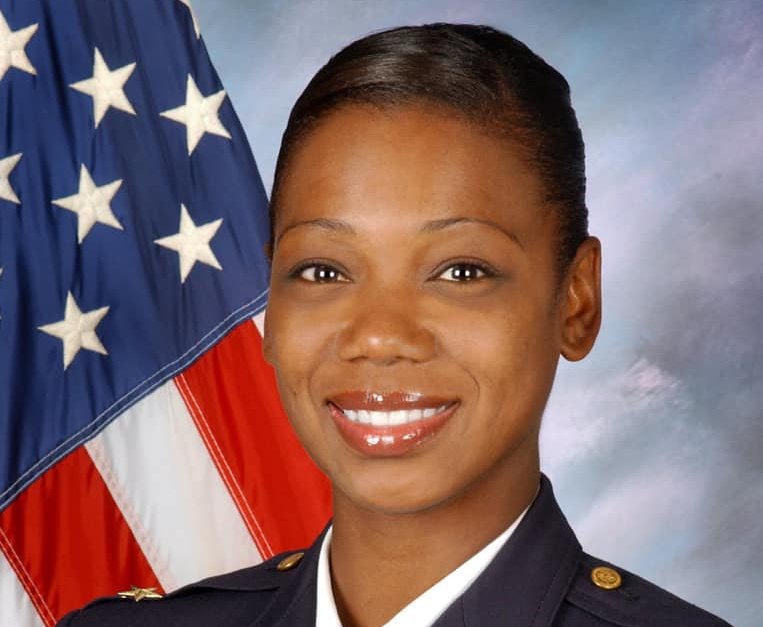 Photograph of Keechant Sewell smiling at the camera in front of a US flag
