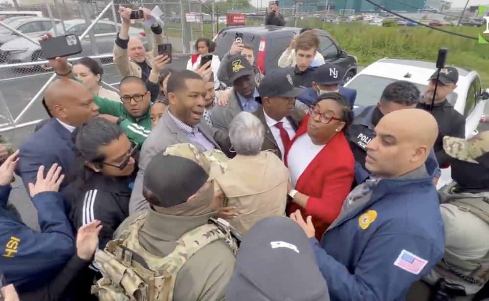 A crowd of people pushing and shoving, some in jackets indicating they're federal agents, outside a detention center.