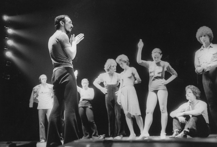 A man stands on a stage in front of several dancers during a rehearsal.