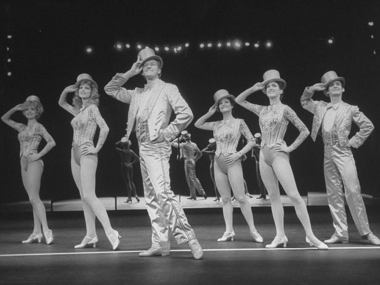 A group of dancers stand in line in matching outfits with top hats.