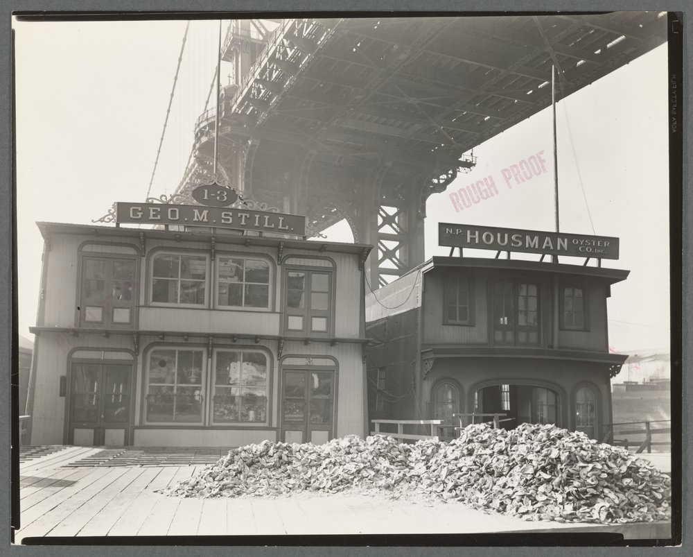 A pile of oysters in front of two old timey taverns in a black and white photo.