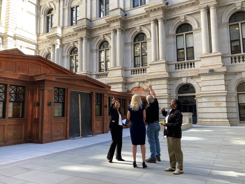 Four people stand inside an interior courtyard with a brass-plated walkway behind them. Two of them are pointing upwards.