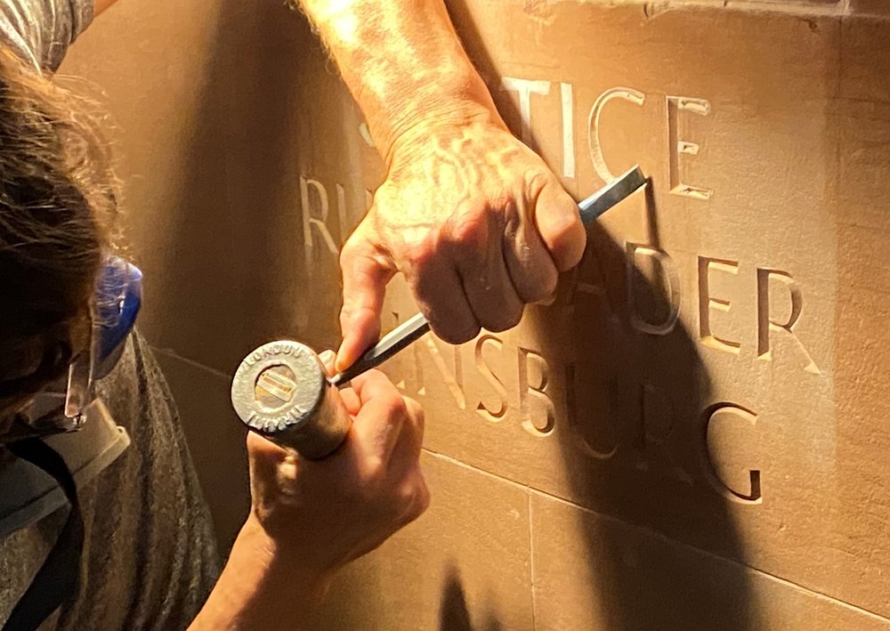 A man wearing a respirator mask carves Ruth Bader Ginsburg's name into a wall using a mallet and chisel.