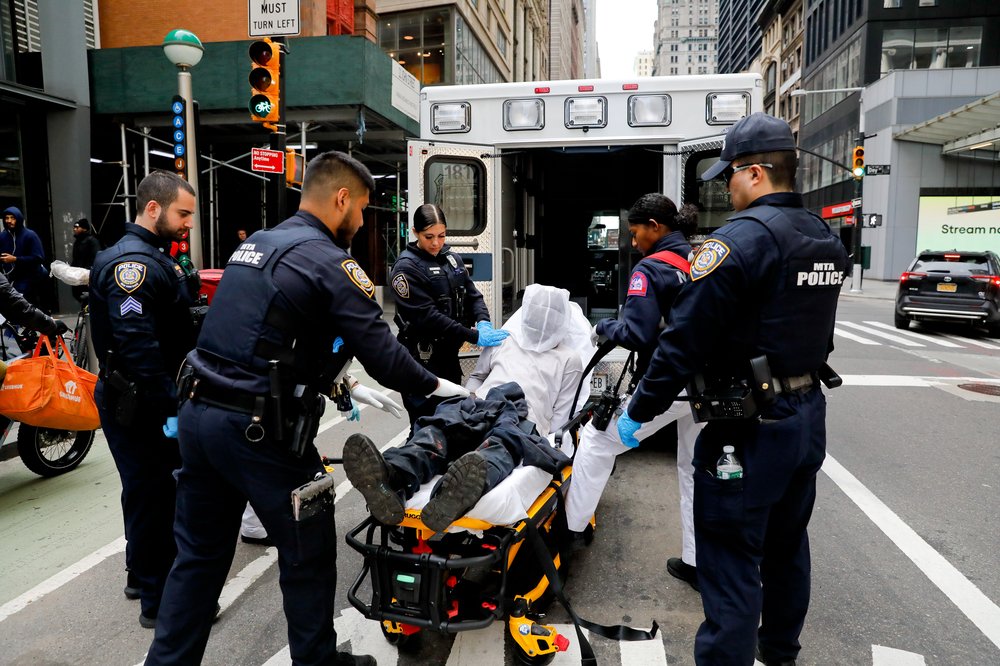 A man with a net over his head is restrained on a gurney and loaded into an ambulance, surrounded by cops.