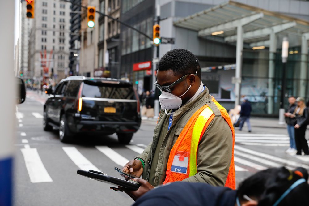 A man wearing a facemask and orange vest holds a clipboard on a Manhattan street.