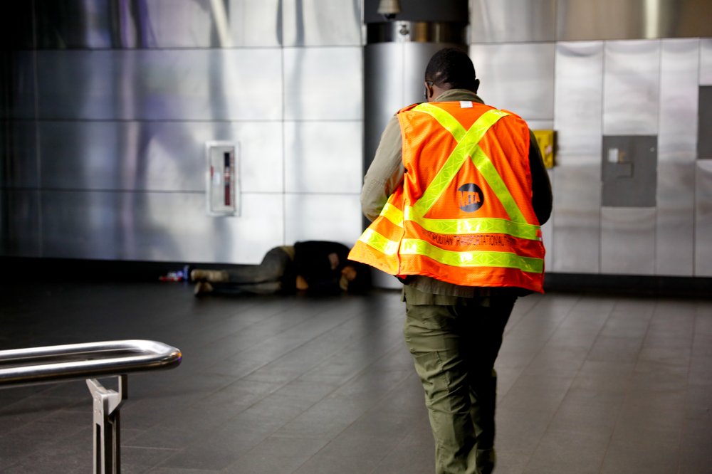 A man an MTA vest tries approaches a man sleeping on the floor of a subway station.