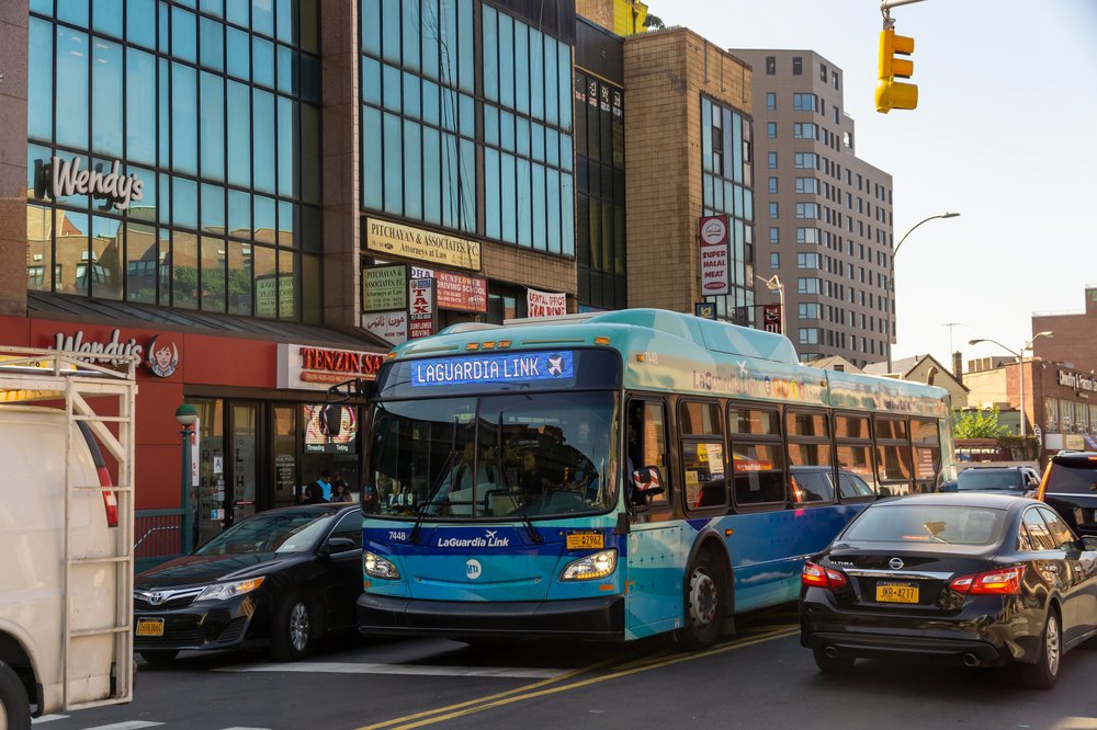 An image of the Q70 bus, which is light blue with dark blue accents and says "Laguardia link" on the digital display in the front, on a Queens Street