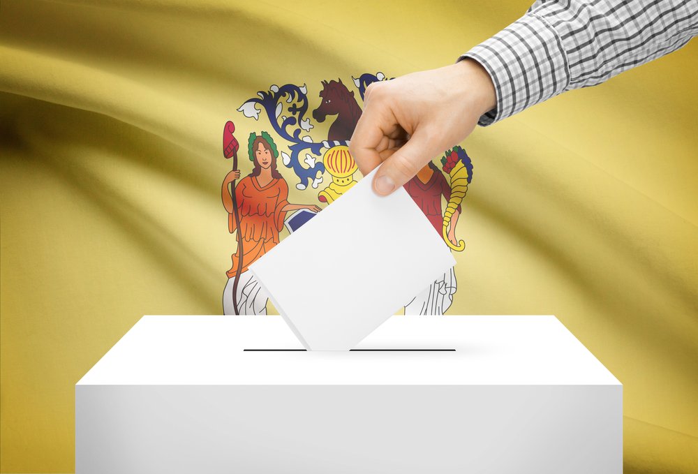 A hand drops a ballot envelope into a box in front of the New Jersey state flag.