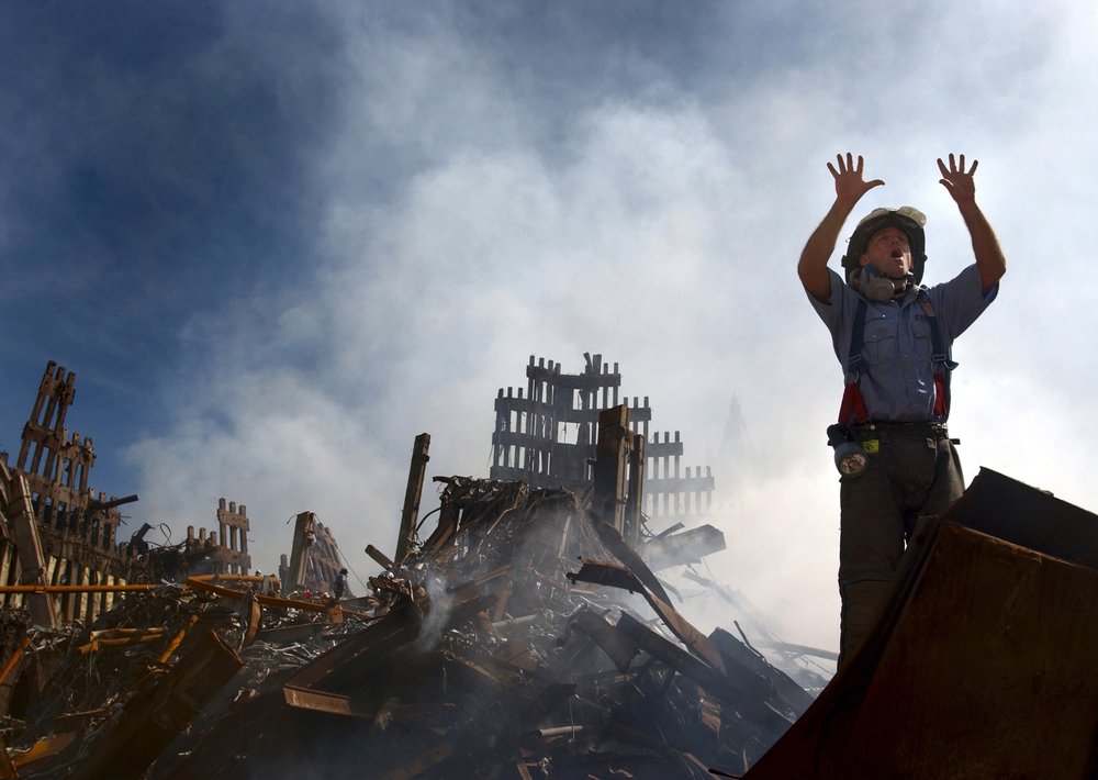 A firefighter without mask and wearing a helmet raises his hands with dusts and debris from the World Trade Center, including the iconic facade, behind him