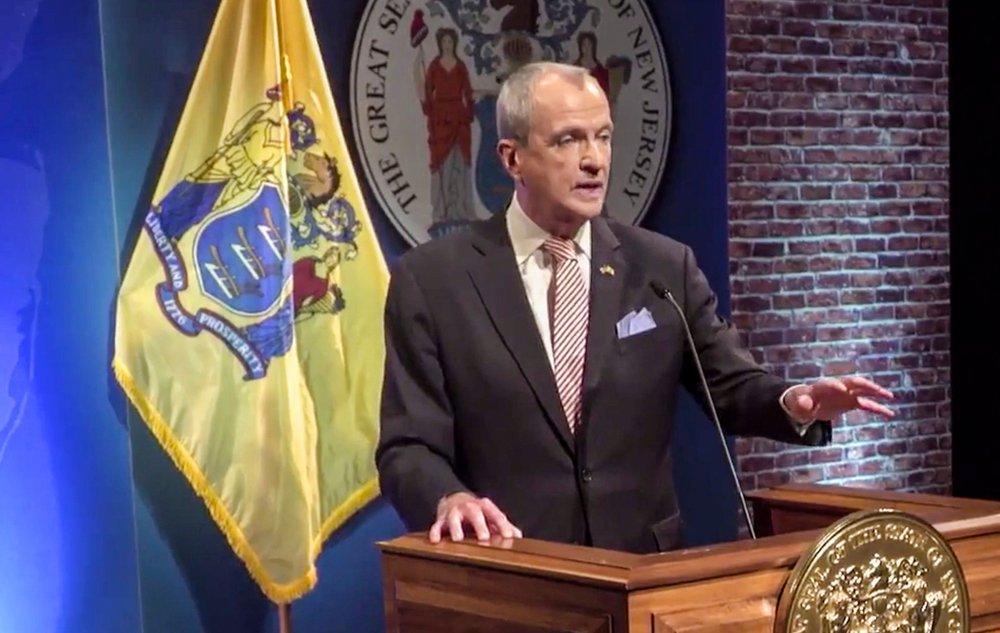 Governor Phil Murphy in a screenshot of his state of the state; he is wearing a suit and standing at a lectern in from of the NJ flag and state seal