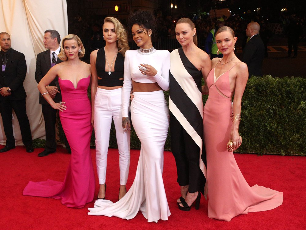 Rihanna is in the center of other stars and fashion designer Stella McCartney. Reese Witherspoon is wearing a strapless slim cut pink gown; Cara Delevigne is wearing a black halter top and slim white pants; Stella McCartney is wearing a black and white one shouldered dress and Kate Bosworth is wearing a pink blush gown.