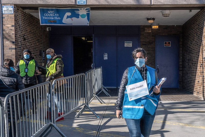 A woman wearing a sign that says "I speak Spanish" "se habla espanol" stands outside Hillcrest High School in Queens where the city runs a vaccination site. Several people stand in line on the left side of the picture.