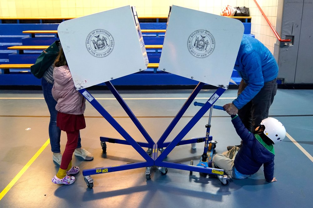 Two people with children vote using New York City voting booths.