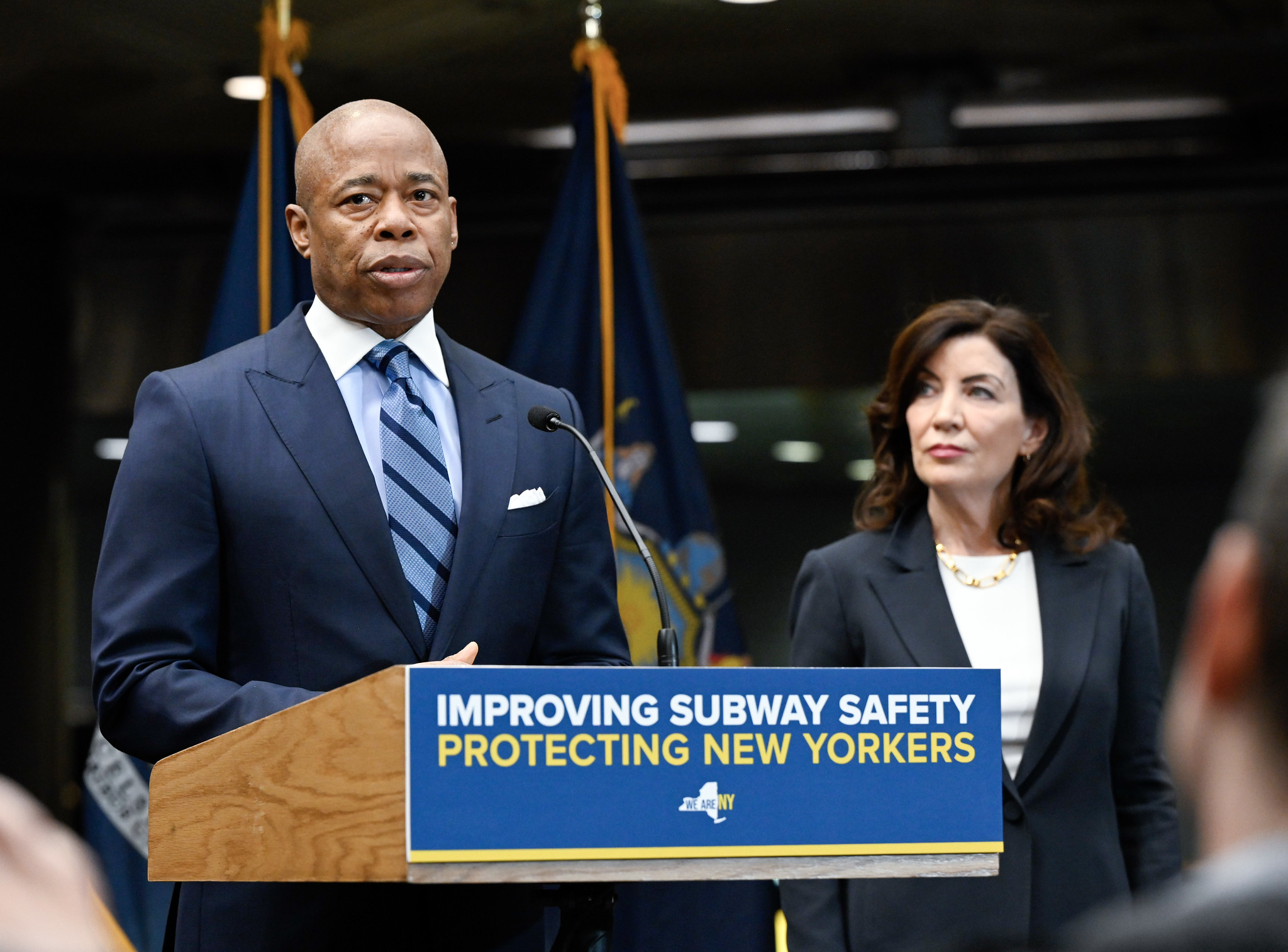 Eric Adams stands at a podium and Kathy Hochul looks at him from behind.