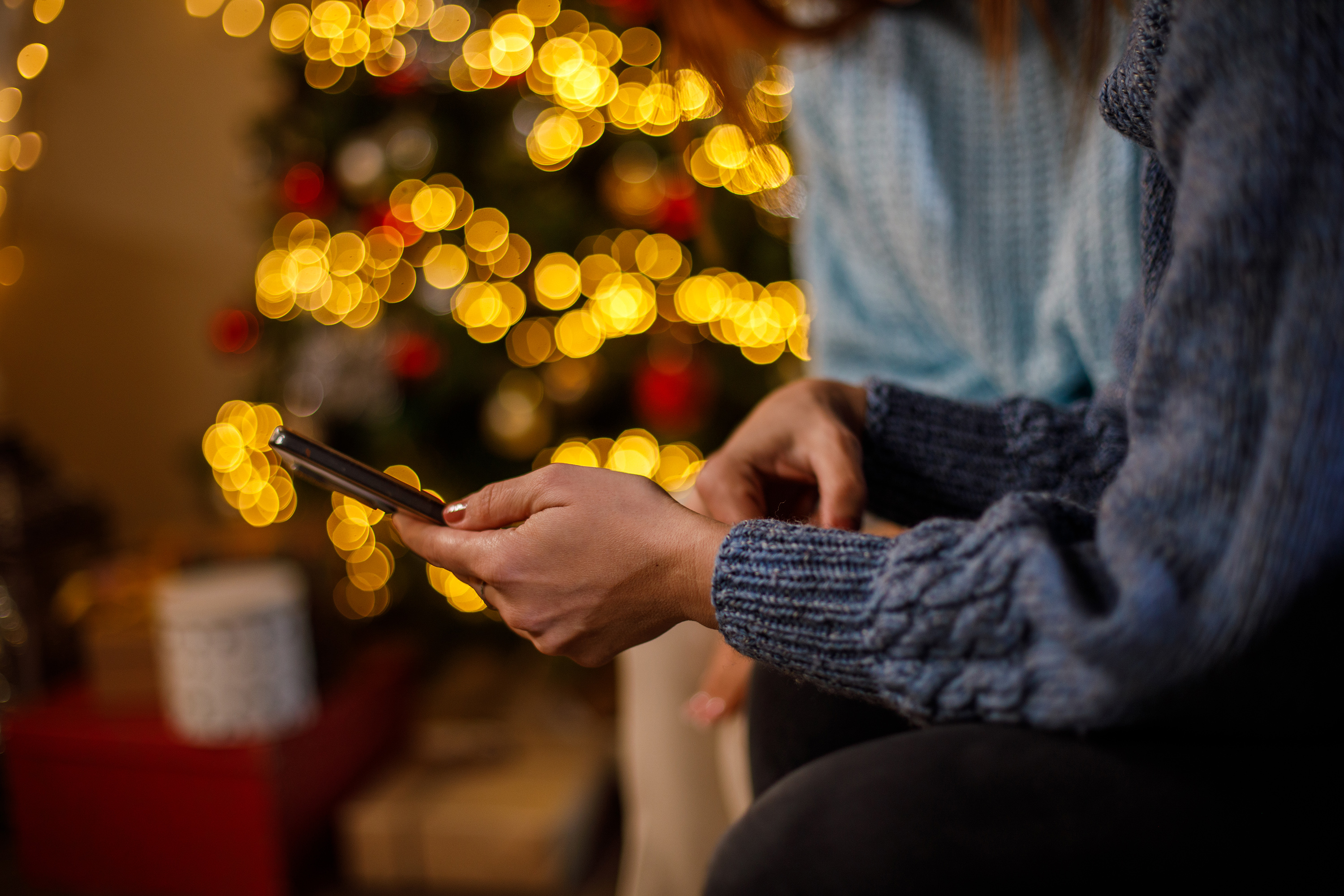 A person's hands is seen using a mobile phone, with a Christmas tree in the background.