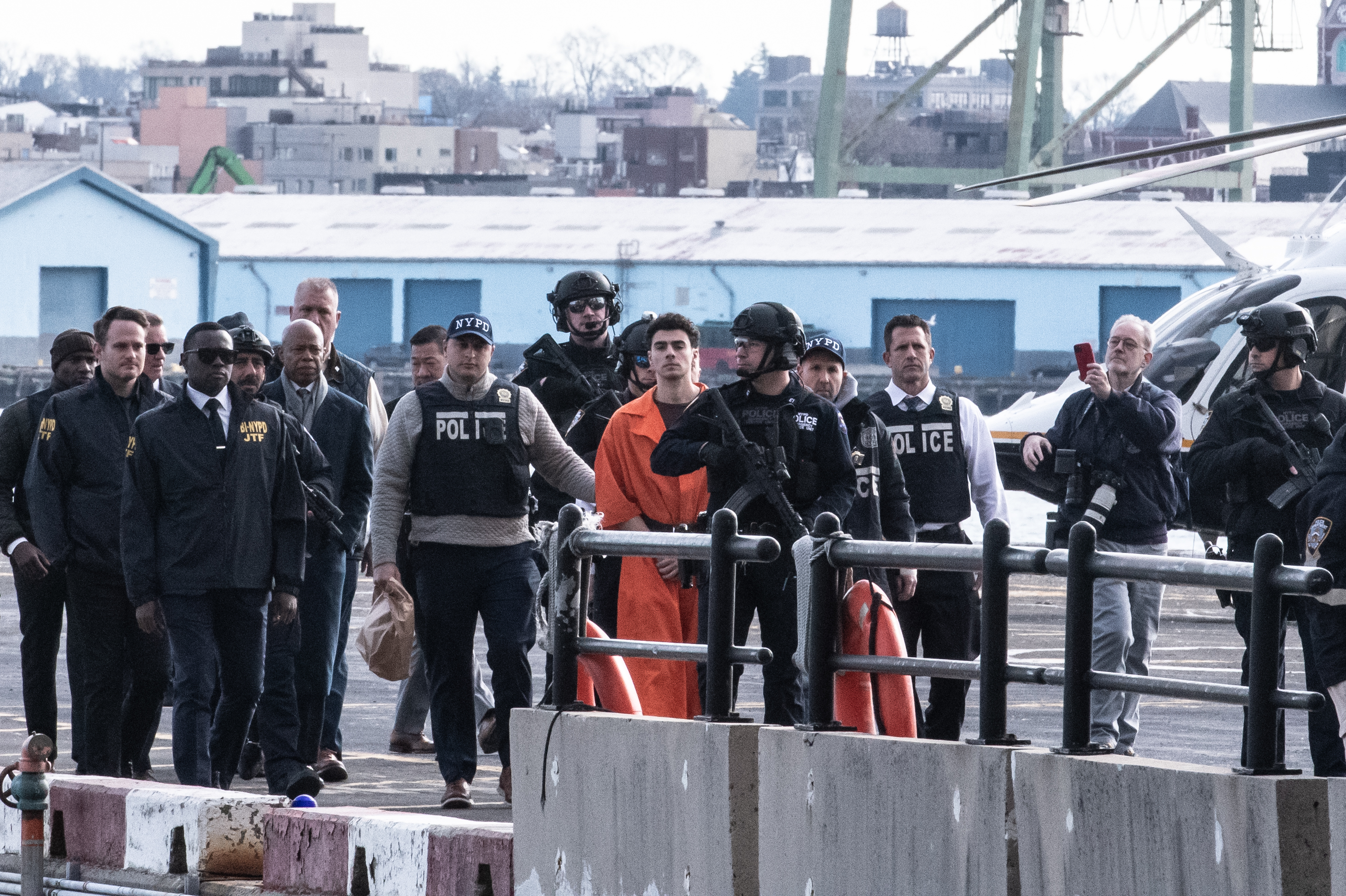 A man in an orange jumpsuit is at a heliport surrounded by uniformed NYPD officers.