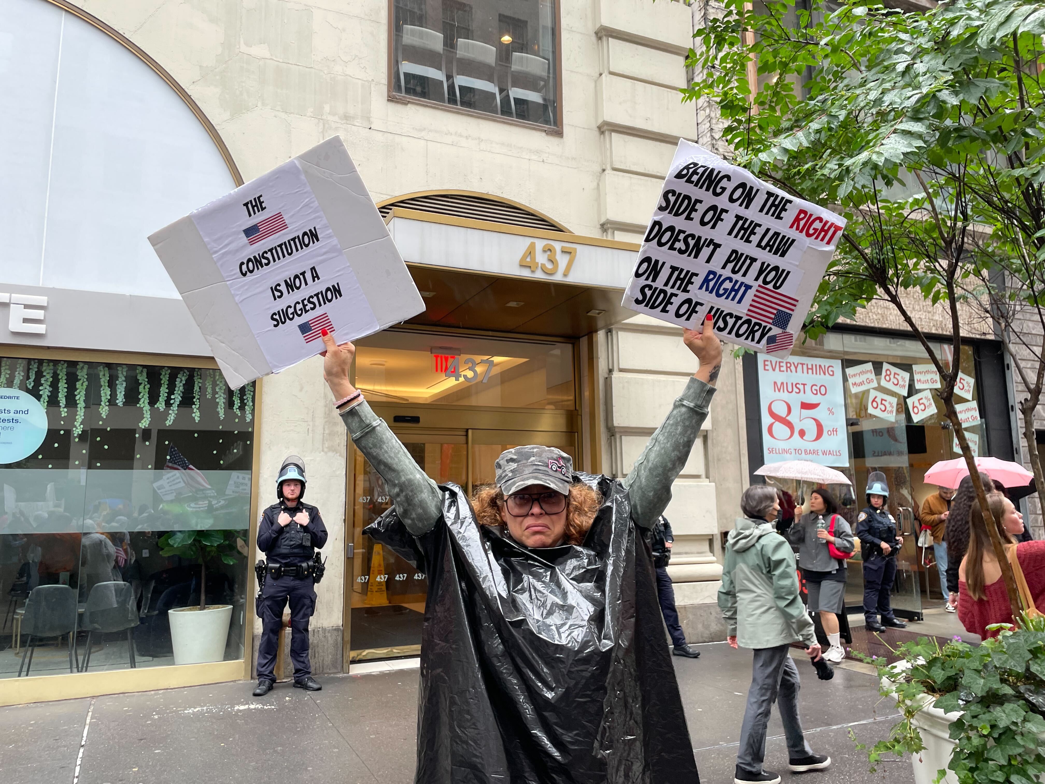 A Latina protester in her 40's or 50's holds signs condemning the Trump Administration's behavior. In the background are NYPD officers in tactical gear and a storefront clearance sign that says 'selling to bare walls'